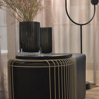 Black ribbed vases with clear bases, set on a sleek black table with gold accents, against soft beige curtains.