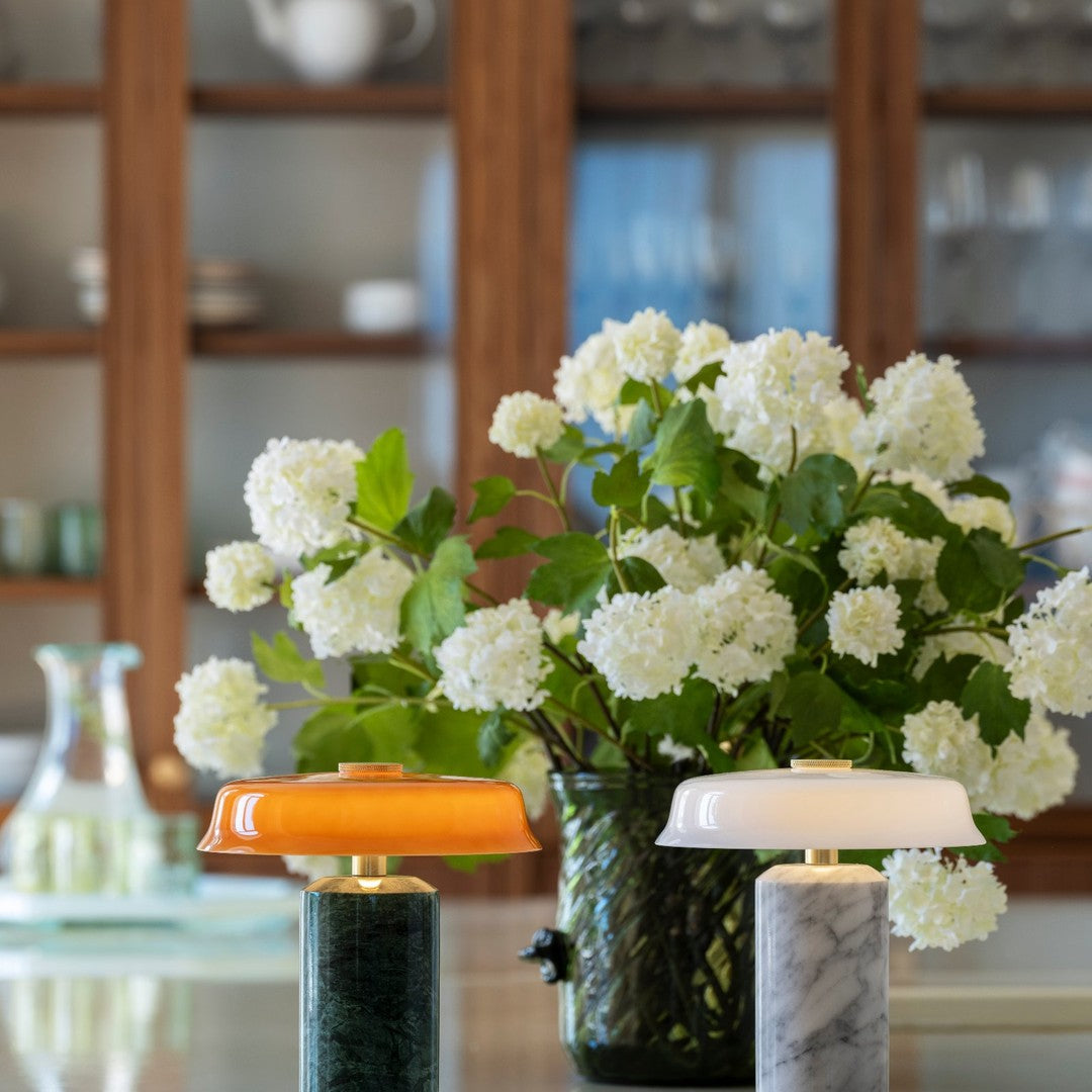 "Orange and white lamps with marble bases, surrounded by lush green foliage and white flowers on a light background."