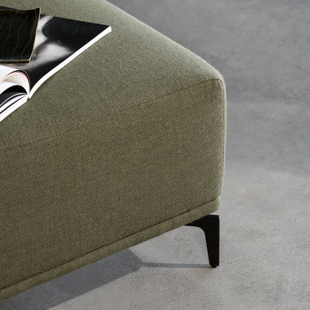 Green textured fabric ottoman with sleek black legs on a gray surface. Modern and stylish design elements.