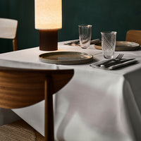White tablecloth with subtle sheen, wooden chairs, decorative glassware, and a warm, textured lamp, against a dark green backdrop.