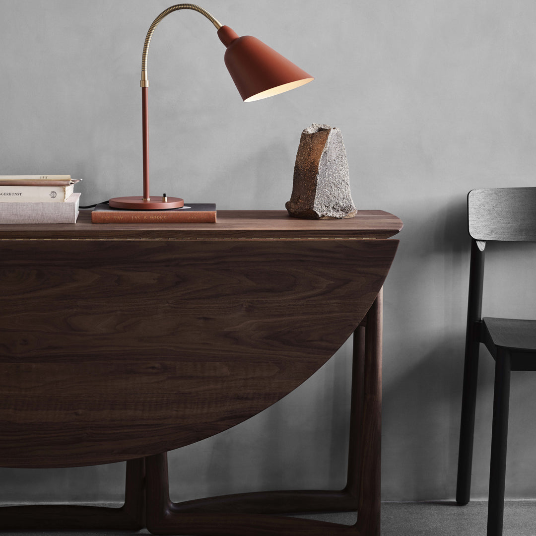 Wooden half-moon table in deep brown with black chair, rust-colored lamp, and grey wall backdrop. Modern minimalist decor.