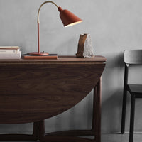 Wooden half-moon table in deep brown with black chair, rust-colored lamp, and grey wall backdrop. Modern minimalist decor.