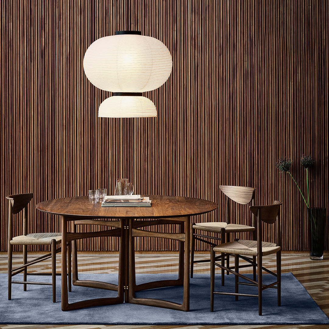 Round wooden dining table with light beige chairs, white pendant lamp, and striped blue rug against a wooden backdrop.