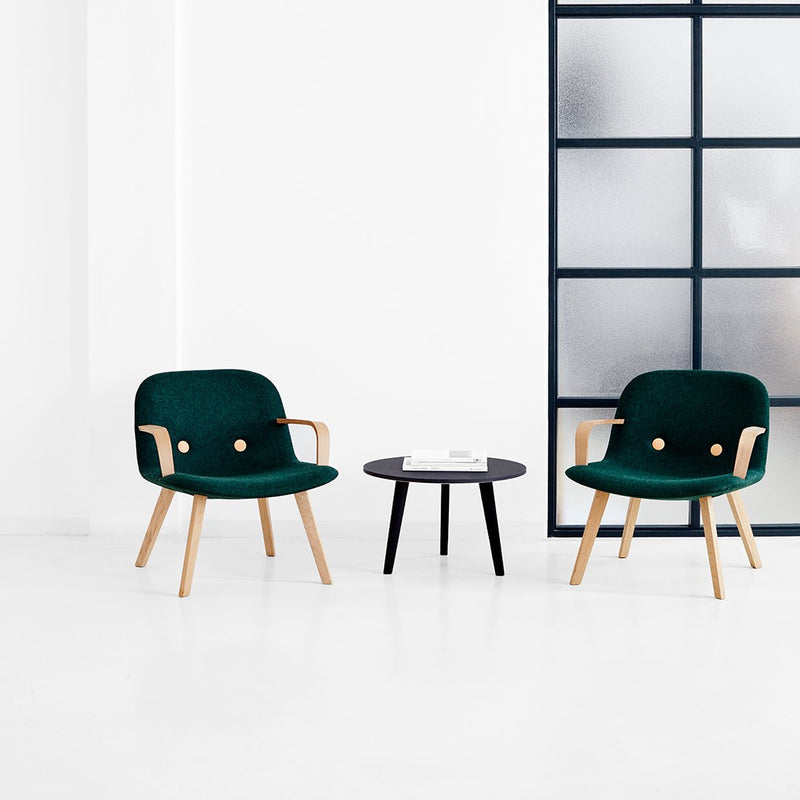 "Two green fabric chairs with wooden arms next to a black round table in a minimalist white room."