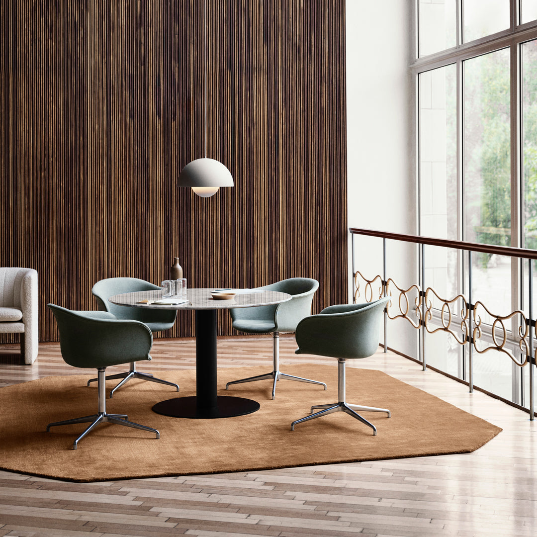 Round table with a sleek black base, surrounded by green chairs on a warm brown hexagonal rug, against a wood panel wall.