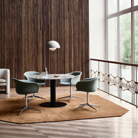 Round table with a sleek black base, surrounded by green chairs on a warm brown hexagonal rug, against a wood panel wall.
