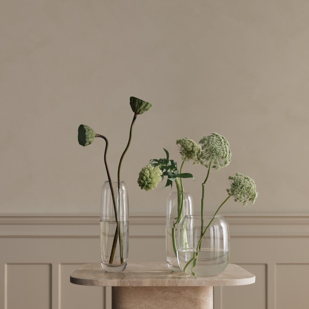Transparent glass vases with green botanical accents on a neutral stone table, featuring natural textures and elegant simplicity.