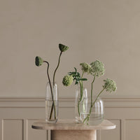 Transparent glass vases with green botanical accents on a neutral stone table, featuring natural textures and elegant simplicity.