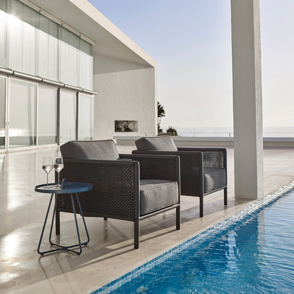 "Two dark gray wicker chairs with plush cushions, paired with a small blue table by a serene poolside setting."