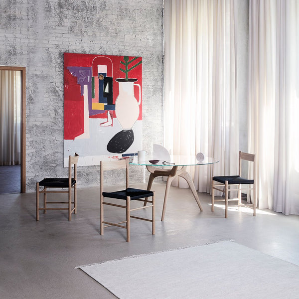 Modern dining room with a colorful abstract wall art, black chair cushions, and light wood accents. Soft neutral tones throughout.