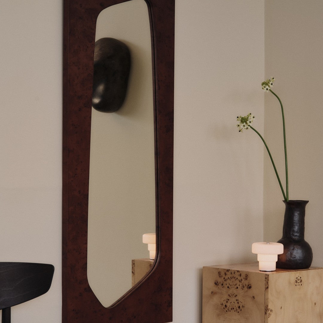 Elegant brown textured frame mirror against a soft beige wall, complemented by a black vase and delicate green stems.