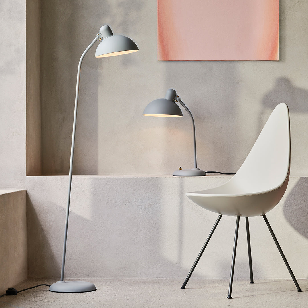 Modern gray floor and desk lamps paired with a white chair and soft pastel wall art against a minimalist backdrop.