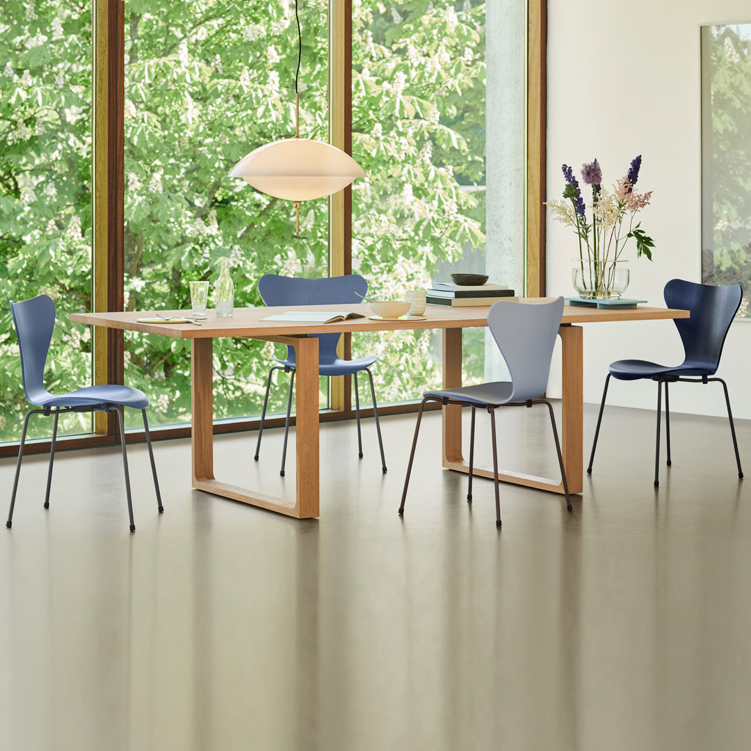 Modern dining room featuring a light wood table, blue chairs, and green outdoor views through large windows.