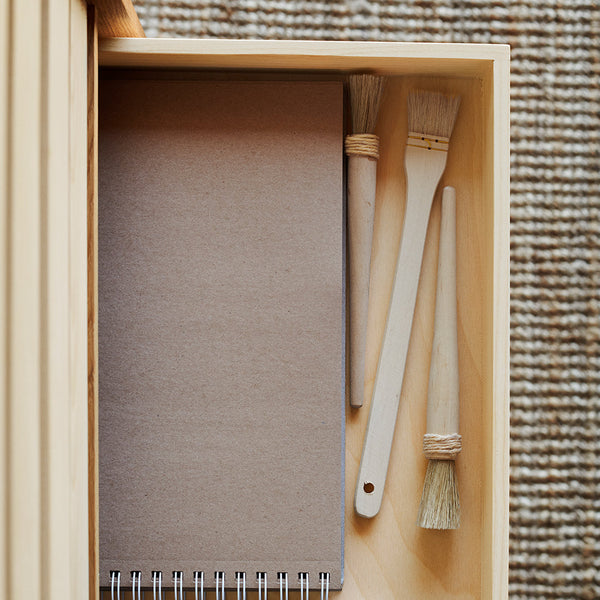 "Neutral brown notebook and wooden brushes inside a light wooden drawer, with a textured woven background."