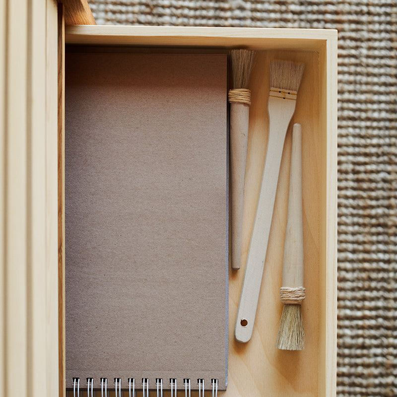 "Neutral brown notebook and wooden brushes inside a light wooden drawer, with a textured woven background."