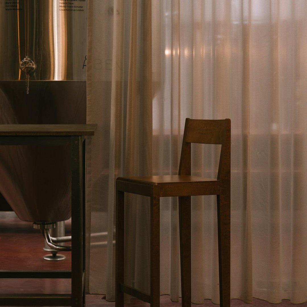 Wooden bar stool against sheer beige curtains, complemented by a warm, muted pink floor. Minimalist, modern design.