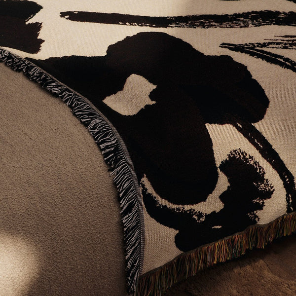 Black and cream abstract pattern woven throw with decorative fringe detail on a beige background.