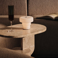 White ribbed lamp and dark green glass on a beige marble table, set against a textured gray backdrop.