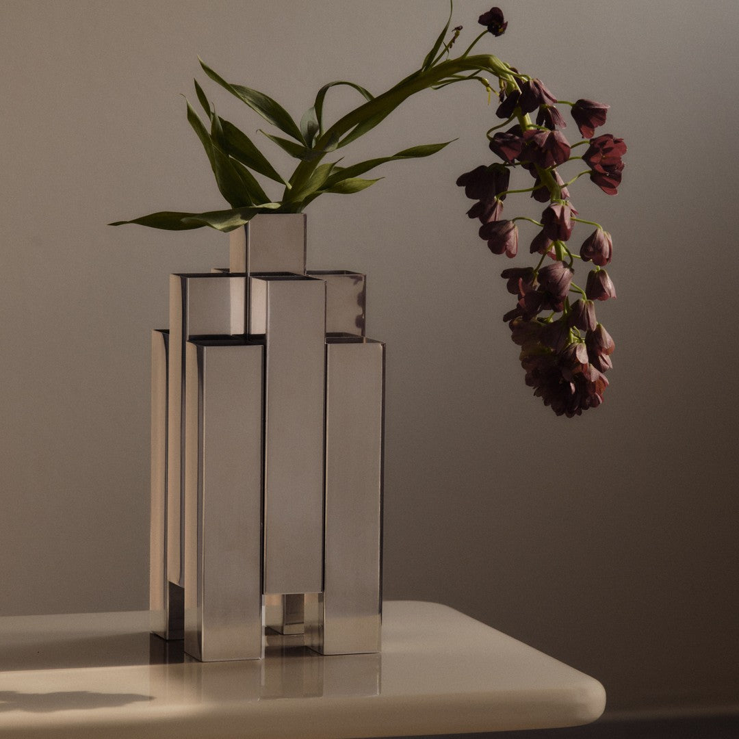 Sleek silver geometric vase holding dark purple flowers with green leaves, set against a neutral background.
