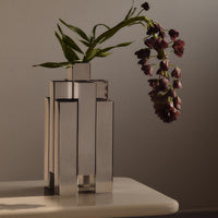 Sleek silver geometric vase holding dark purple flowers with green leaves, set against a neutral background.