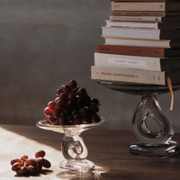 "Transparent glass bowl holding dark red grapes, stacked books in earthy tones, soft shadows on a wooden surface."