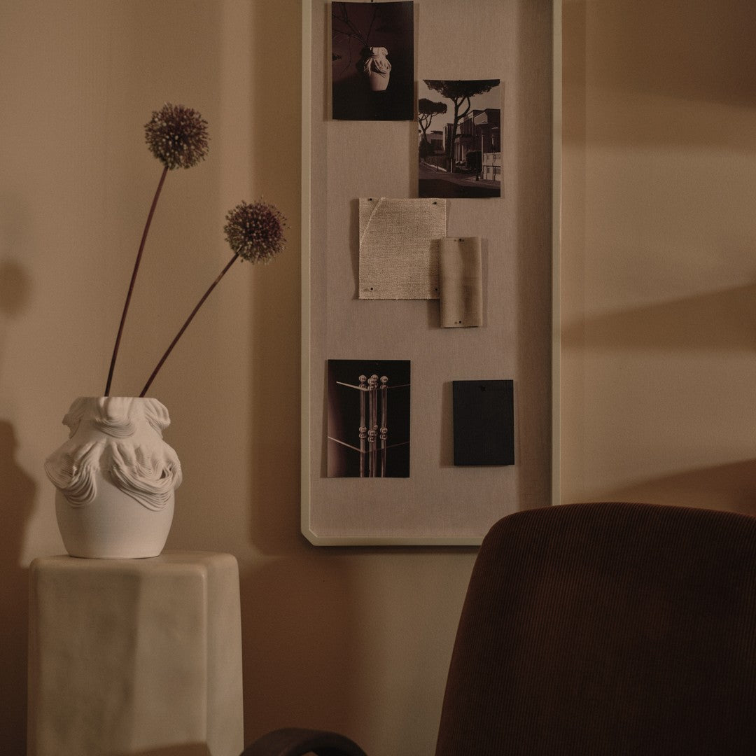 Elegant white vase with textured design, paired with dark prints, neutral beige wall, and warm brown chair.