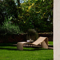 Beige modern lounge chair on lush green grass, surrounded by leafy trees and a sleek black side table.