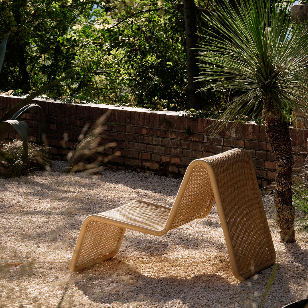 Sleek tan lounge chair with a modern wavy design, set against a lush green garden backdrop.