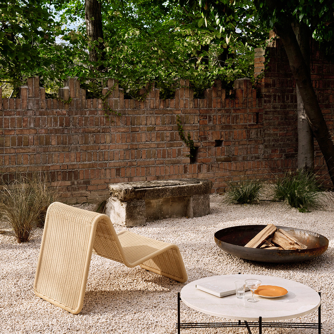 Natural woven lounge chair in beige, surrounded by a neutral gravel setting and rustic brick wall accents.