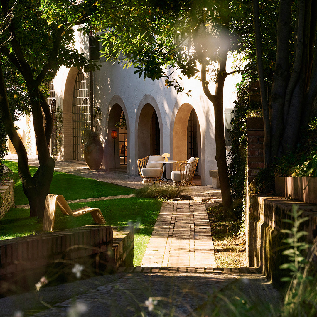 "Sunlit outdoor pathway with vibrant greenery, rustic stone, and beige furniture, creating a serene garden ambiance."