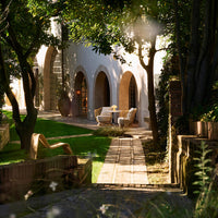 "Sunlit outdoor pathway with vibrant greenery, rustic stone, and beige furniture, creating a serene garden ambiance."
