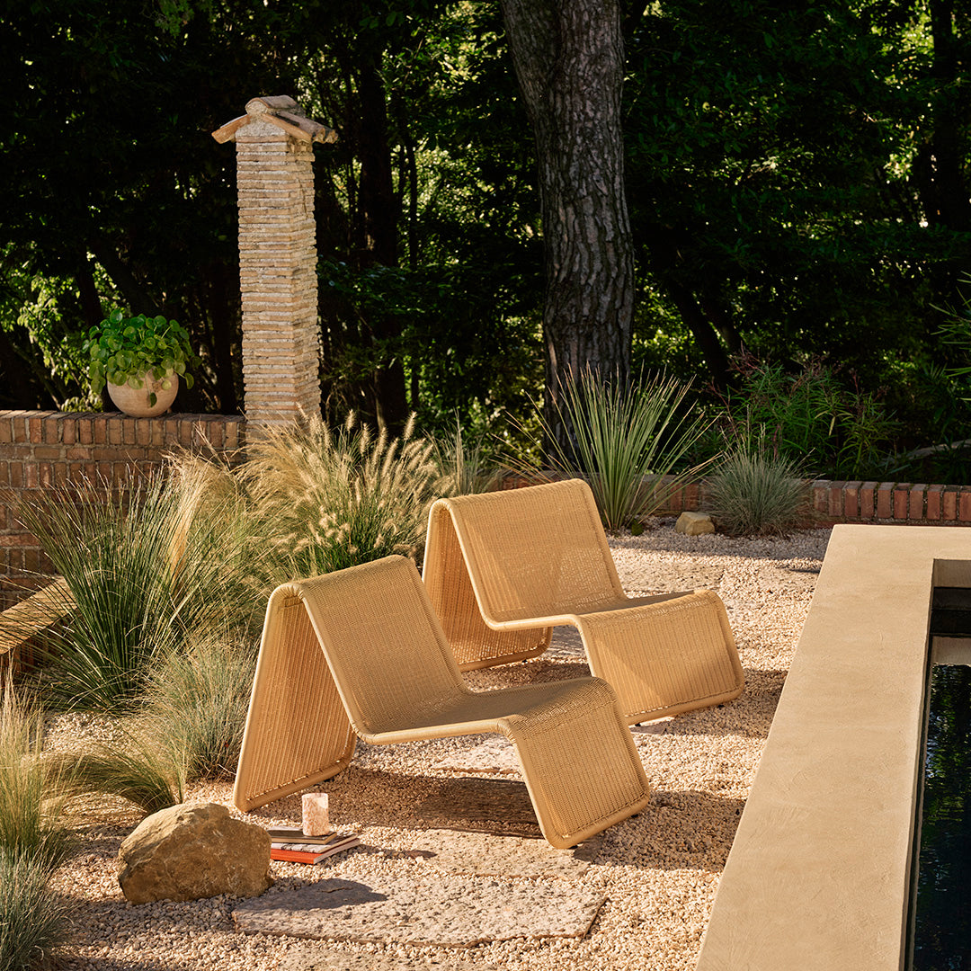 "Minimalistic tan folding chairs with clean lines, set against a natural outdoor landscape of greenery and stone."