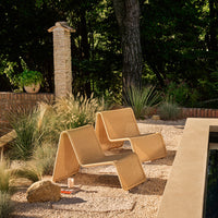"Minimalistic tan folding chairs with clean lines, set against a natural outdoor landscape of greenery and stone."