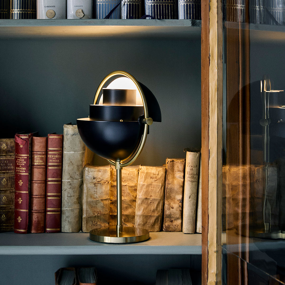 Black two-toned lamp with a gold base, positioned among vintage books on a dark shelf. Elegant and modern design.