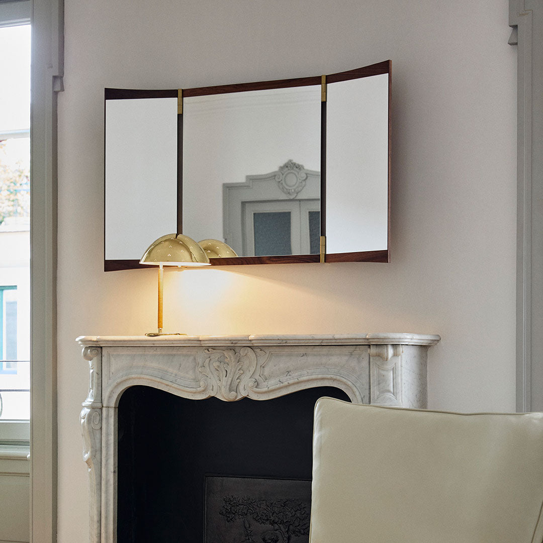 Elegant tri-fold mirror with a dark wood frame and gold accents against a soft, neutral wall, reflecting natural light.