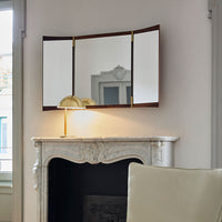 Elegant tri-fold mirror with a dark wood frame and gold accents against a soft, neutral wall, reflecting natural light.
