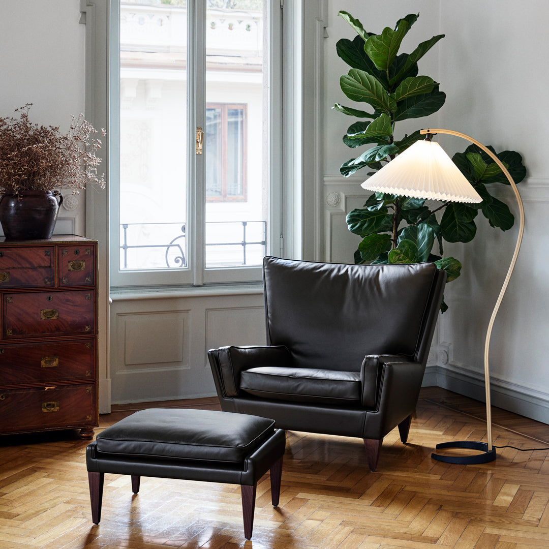 Black leather armchair and ottoman with natural wood floor, accented by a cream-colored lamp and lush green plant.