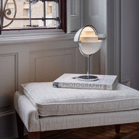 Modern round lamp in white with chrome accents, placed on a gray upholstered ottoman beside a photography book.