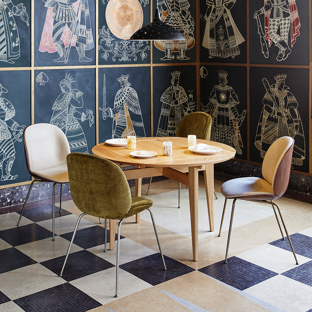 "Round wooden table with colorful velvet chairs in olive, cream, and brown, set against patterned black and white flooring."