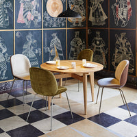 "Round wooden table with colorful velvet chairs in olive, cream, and brown, set against patterned black and white flooring."