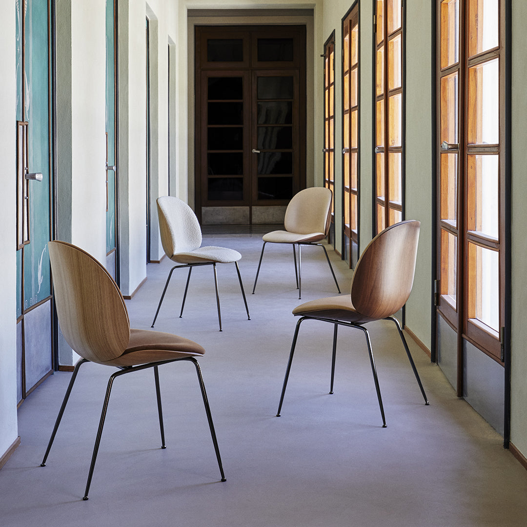 Four minimalist chairs with soft beige upholstery and black metal legs arranged in a spacious hallway with natural light.