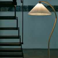 White pleated lampshade with a sleek, curved metallic base, set against a neutral background and dark staircase.