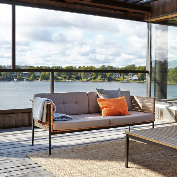 Modern outdoor sofa in gray upholstery, accented with an orange pillow and light blue throw, set against a serene lakeside backdrop.