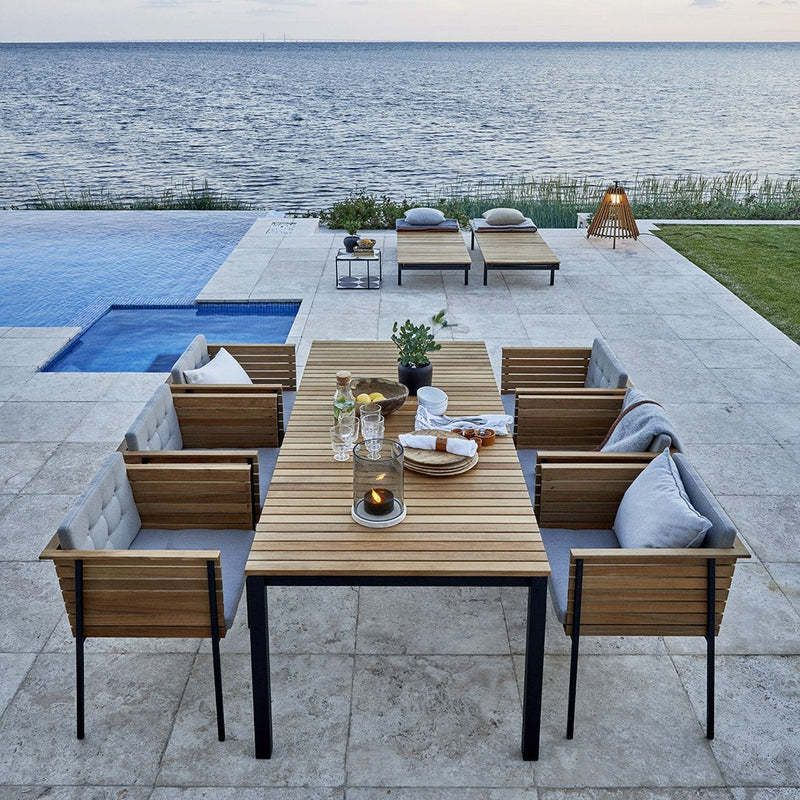 Elegant outdoor dining set featuring wooden accents, gray cushions, and a neutral stone patio by a serene blue pool.