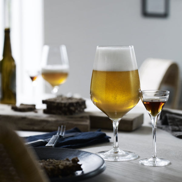 Golden beer with frothy white head, served in elegant glassware, alongside a small shot glass. Neutral table settings.
