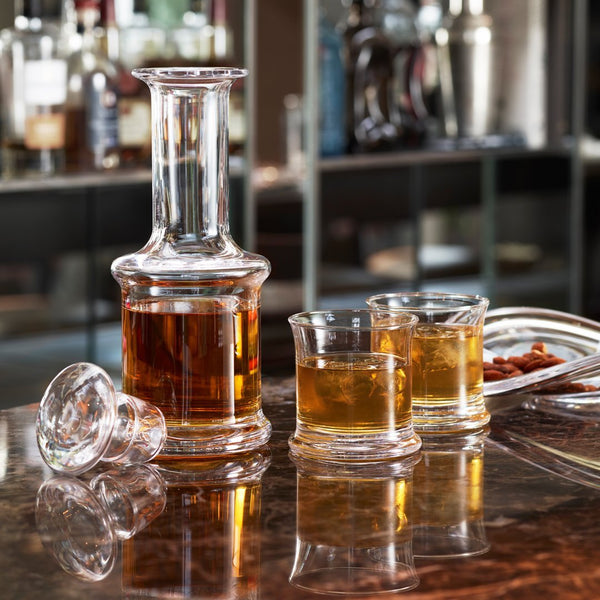 Clear glass decanter and whiskey tumblers with amber liquid, reflecting light on a dark marble surface.