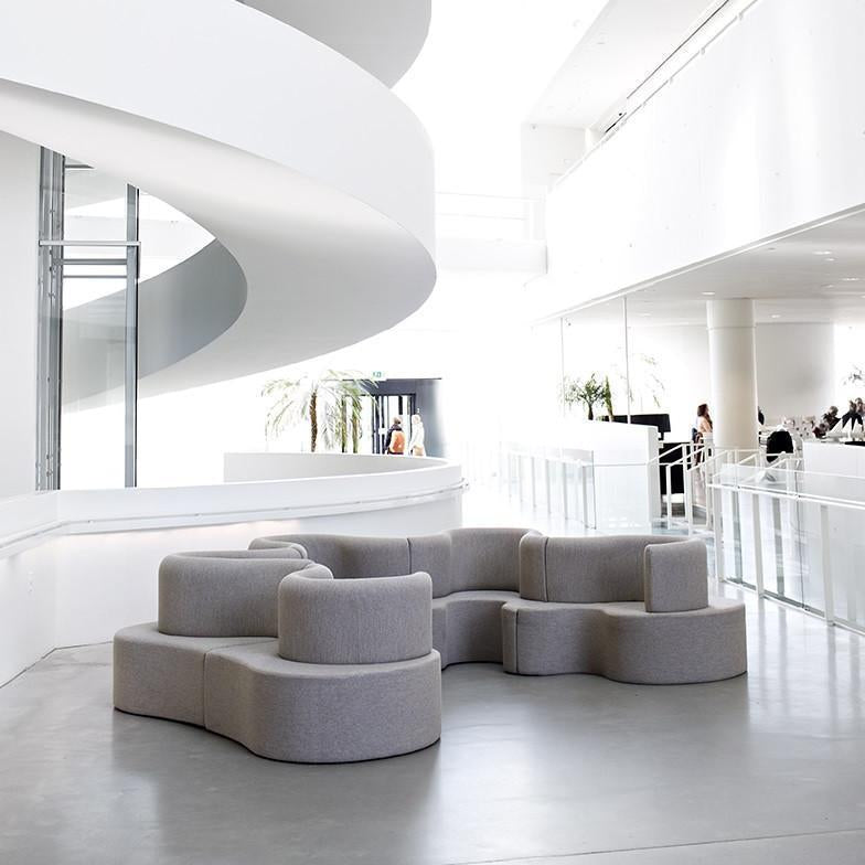 Curved gray sofa with textured fabric, set against a bright white backdrop and smooth concrete floor. Modern minimalist design.