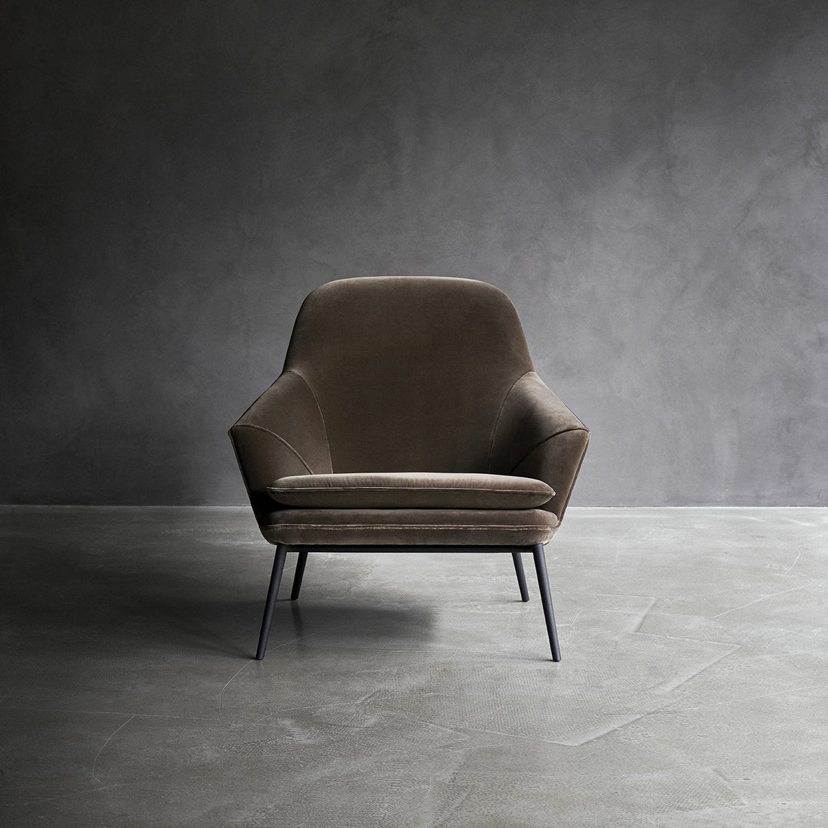 Brown velvet accent chair with smooth, modern lines and sleek black legs, set against a minimalist gray backdrop.