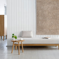 Neutral beige wall hanging, soft gray couch, light wooden table, and green plant accents against a white wall.