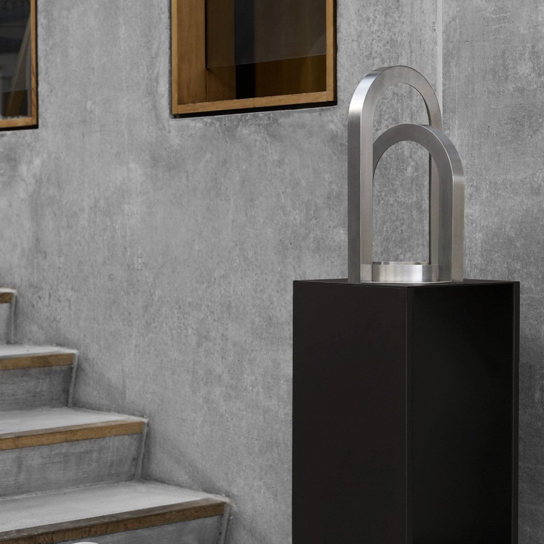 Modern black pedestal with silver arch design, set against a textured gray wall and wooden staircase.